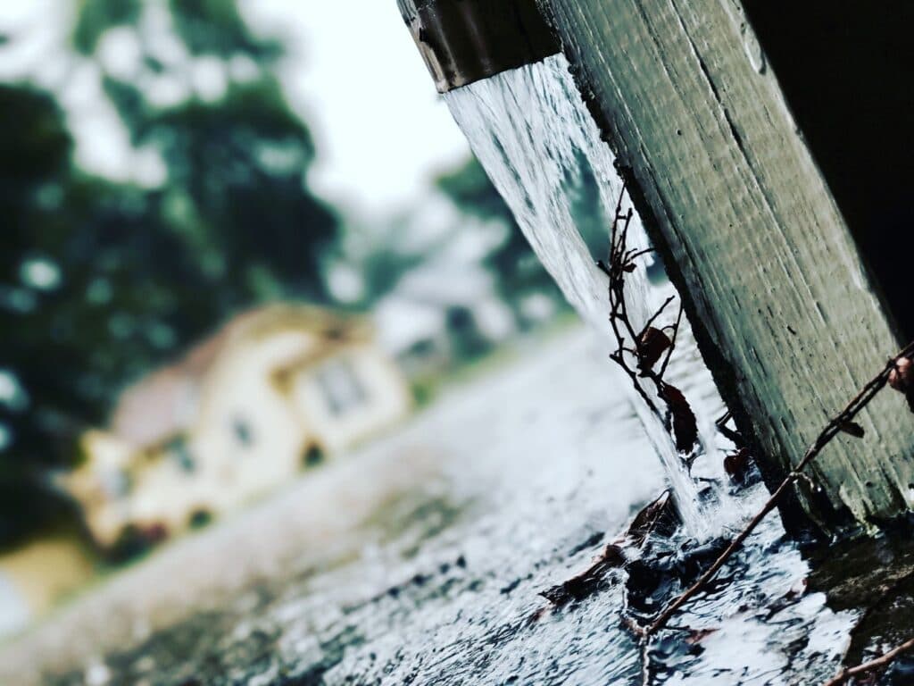 Rainwater pouring hard from a downspout directly onto pavement beside a home's foundation