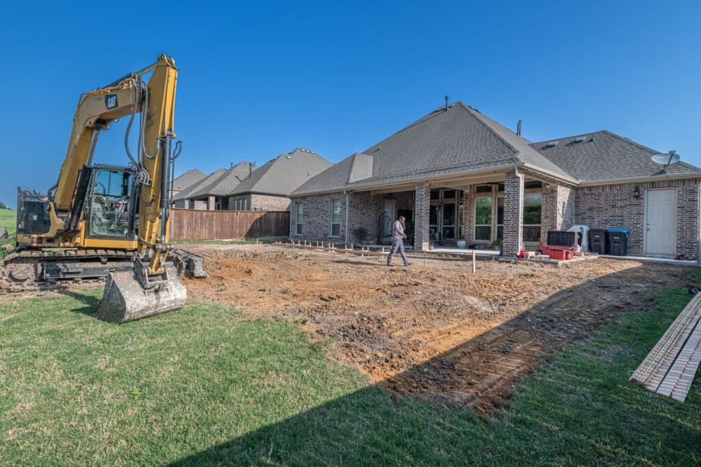 Excavator and staked layout marking the patio base excavation before an outdoor kitchen build
