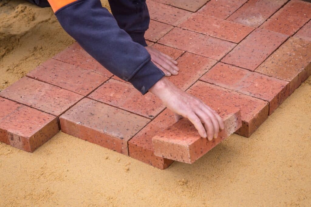 Worker's hands setting an individual brick paver into a sand bed during a driveway repair job